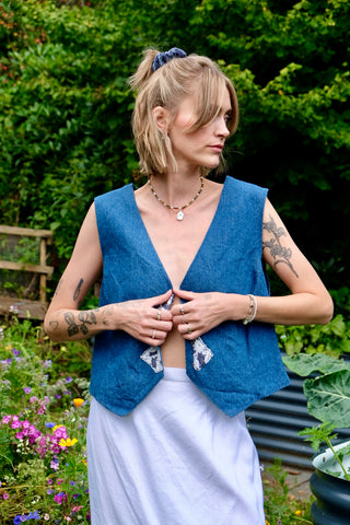 Kurz geschnittene 90s Handmade Jeansweste in mittelblauer Waschung mit Hakenverschlüssen und Details aus weißer Spitze sowie Bändchen zum Binden am Rücken – wir empfehlen die Weste einer S – ein absolutes Highlight