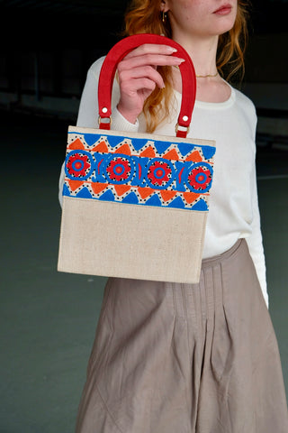 Cremeweiße/Beige Preloved (Secondhand) Handmade Handtasche im Vintage Stil von der Fair Trade Marke Aarong aus Bangladesch, die ländliche Kunsthandwerker unterstützt. Die Marke ist eine soziale Initiative der gemeinnützigen Organisation&nbsp;BRAC International. Die Tasche hat eine hübsche quadratische Form mit roten Henkeln und ist mit tollen Stickereien in Blau, Orange und Rot sowie kleinen schwarzen Pailletten verziert. Mit einem kleinen Innenfach, das mit einem Reißverschluss ausgestattet ist.