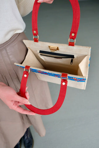 Cremeweiße/Beige Preloved (Secondhand) Handmade Handtasche im Vintage Stil von der Fair Trade Marke Aarong aus Bangladesch, die ländliche Kunsthandwerker unterstützt. Die Marke ist eine soziale Initiative der gemeinnützigen Organisation&nbsp;BRAC International. Die Tasche hat eine hübsche quadratische Form mit roten Henkeln und ist mit tollen Stickereien in Blau, Orange und Rot sowie kleinen schwarzen Pailletten verziert. Mit einem kleinen Innenfach, das mit einem Reißverschluss ausgestattet ist.