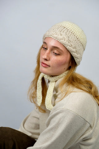 Handmade Bonnet aus Schurwolle-Mix-Garn in Cremeweiß mit Zopfstrickbündchen in Beige und Bindebändern.