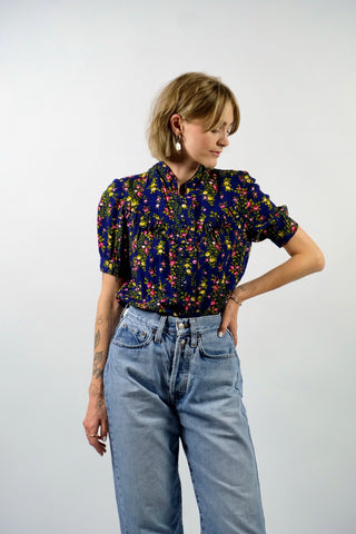 Dunkelblaue Handmade 70s Bluse mit kleinem Stehkragen, leichten Puffärmeln und Blümchenmuster in Grün, Gelb, Rot, Pink und Weiß – wir empfehlen die Bluse einer XXS