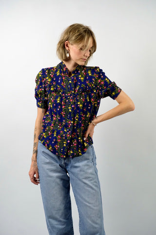 Dunkelblaue Handmade 70s Bluse mit kleinem Stehkragen, leichten Puffärmeln und Blümchenmuster in Grün, Gelb, Rot, Pink und Weiß – wir empfehlen die Bluse einer XXS